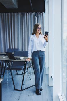 Photo of a successful young woman in a shirt smiling and working on a laptop while talking on the phone in a modern office with large windows. Remote work with coffee