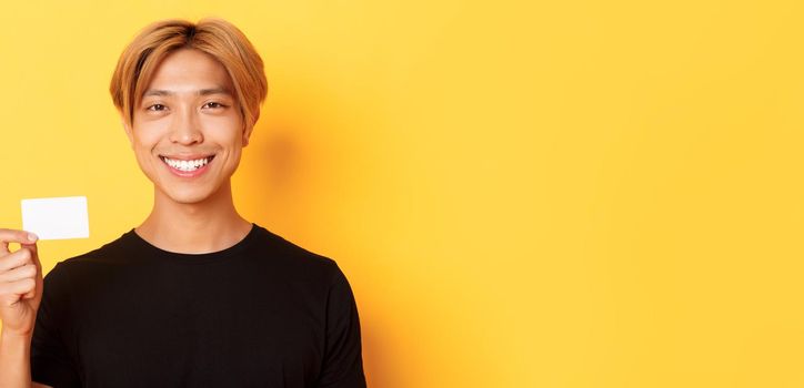 Close-up of handsome pleased asian guy with fair hair, smiling happy and showing credit card over yellow background.