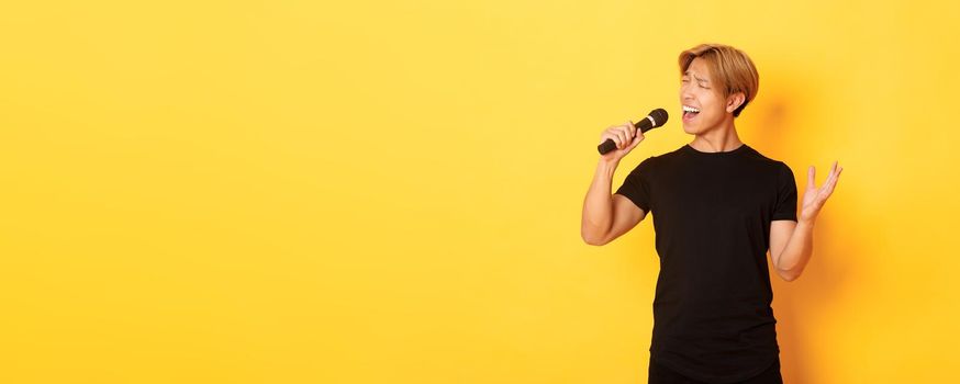 Carefree handsome asian guy, korean singer singing into microphone with passion, standing yellow background.