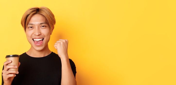 Close-up of energized handsome asian guy fist pump joyfully while drinking coffee, smiling excited over yellow background.