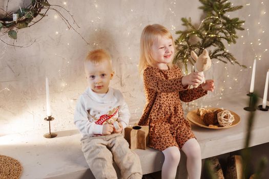 Little brother and sister play on Christmas eve in a beautiful house decorated for the New Year holidays. Children are playing with a Christmas gift. Scandinavian-style interior with live fir trees and a wooden staircase.