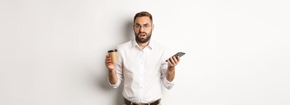 Image of man drinking coffee, feeling confused about strange message on mobile phone, standing over white background.