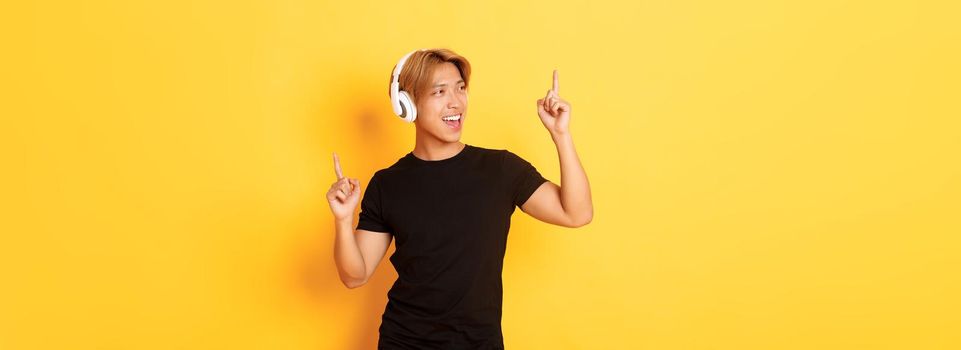 Joyful handsome asian guy with blond hair, singing along and dancing as listening to music in wireless headphones, standing yellow background.