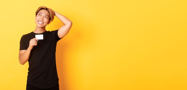 Thoughtful smiling asian guy thinking while showing credit card, looking upper left corner dreamy, yellow background.