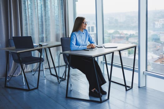 A beautiful woman in glasses sits at a table in the office and works at a laptop. Corporate work in the office. Modern woman workplace manager in classic clothes