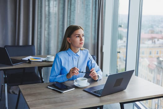 Adult smiling female manager in glasses and formal shirt sits at a desk and looks at a laptop. Modern, spacious, bright office with a large window.