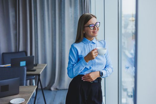 Image of a happy young woman in a classic shirt and glasses smiling and drinking coffee while standing by the window in a modern office with large windows. Remote work