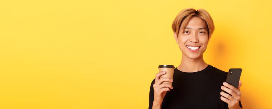 Close-up of handsome young asian guy smiling happy, using smartphone and drinking takeaway coffee, standing over yellow background.