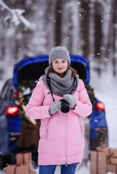 A woman in a winter snow-covered forest in the trunk of a car decorated with Christmas decor. A female photographer holds a camera in her hands.