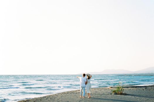 Man shows the sea with his finger to the woman, standing on the beach. Back view. High quality photo