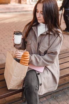 A stylish brunette woman walks around the autumn city. The brunette is sitting on a bench outside with a glass of coffee and a phone, holding a craft bag with a baguette.