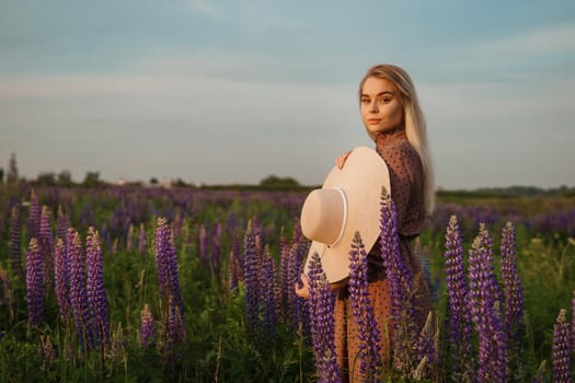 A beautiful woman in a straw hat walks in a field with purple flowers. A walk in nature in the lupin field.