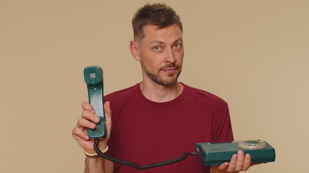 Hey you, call me back. Handsome young man in red t-shirt talking on wired vintage telephone of 80s, says hey you call me back. Call center. Adult stylish male guy on beige studio background indoor