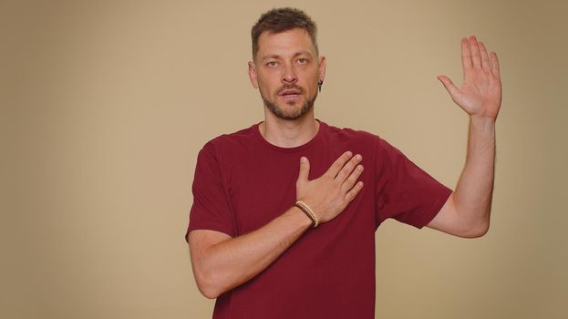 I swear to be honest. Sincere responsible handsome young man raising hand to take oath, promising to be honest and to tell truth, keeping hand on chest. Adult stylish guy isolated on beige background