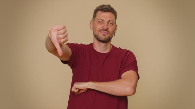 Upset bearded handsome young man showing thumbs down sign gesture, expressing discontent, disapproval, dissatisfied, dislike. Adult stylish male guy isolated alone on beige studio background indoors