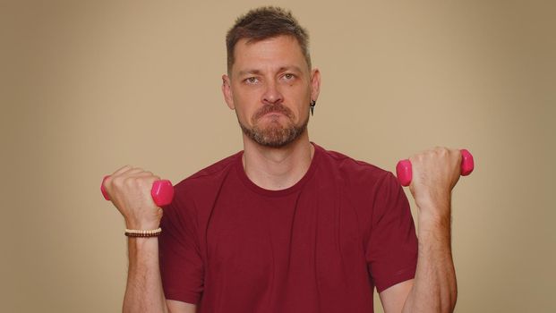 Handsome young man in red t-shirt working out, pumping up arm muscles lifting pink gym dumbbells, practicing pilates, healthcare. Adult stylish guy on beige studio wall background. Sport motivation