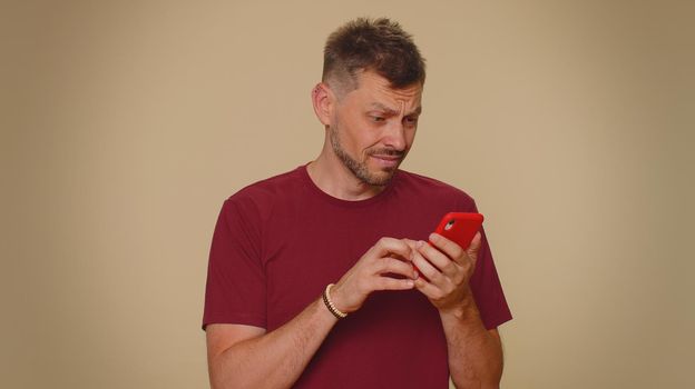 Sad young man in red t-shirt use smartphone typing browsing, loses becoming surprised sudden lottery results, bad fortune, loss. Adult stylish guy isolated on beige studio wall background indoors