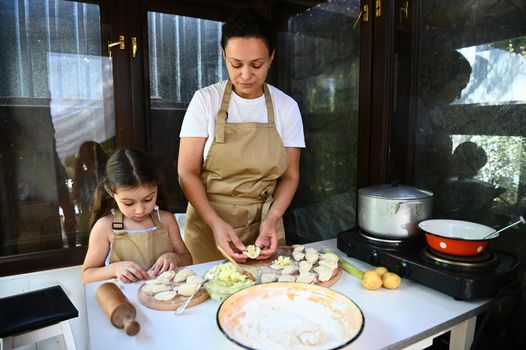 Loving young mother and her adorable little daughter in chef's aprons, enjoy making homemade dumplings together, rolling out dough and stuffing mashed potatoes, standing at table in a country kitchen