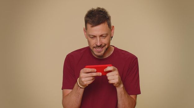 Worried funny young man in red t-shirt enthusiastically playing racing or shooter video games on smartphone. Adult stylish guy using mobile phone gadget app on beige studio background indoors