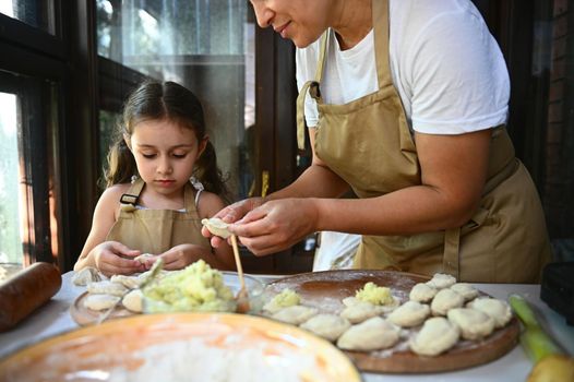 Pretty woman in white t-shirt and beige chef's apron, a loving mother showing her cute daughter how to make homemade dumplings, teaching her how to cook Varenyky according to traditional family recipe