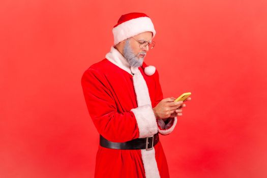 Serious elderly man with gray beard in santa claus costume using smart phone with concentrated facial expression, browsing internet, social network. Indoor studio shot isolated on red background.