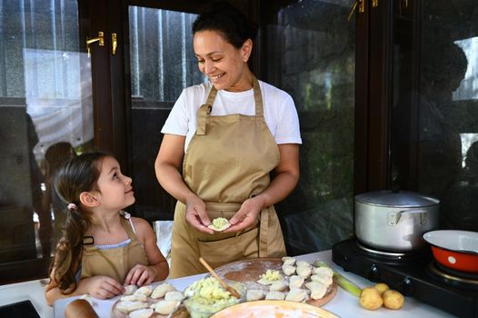 Charming African American woman in chef's apron admiring her cute child, a baby girl who is learning to cook dumplings or vareniki in a summer village kitchen. Mother and daughter baking in kitchen