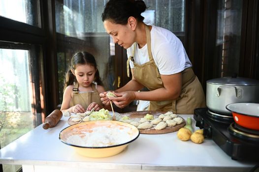 Loving mother in beige chef's apron, stands by a table with dough, teaches her adorable little daughter how to make dumplings, having fun together in the country kitchen. Vareniki Pelmeni Ravioli.