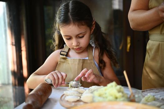 Adorable Caucasian little girl, child cook putting mashed potatoes on a rolled out dough and enjoying cooking dumplings with her loving mother . Pretty little girl learns making Ukrainian vareniki