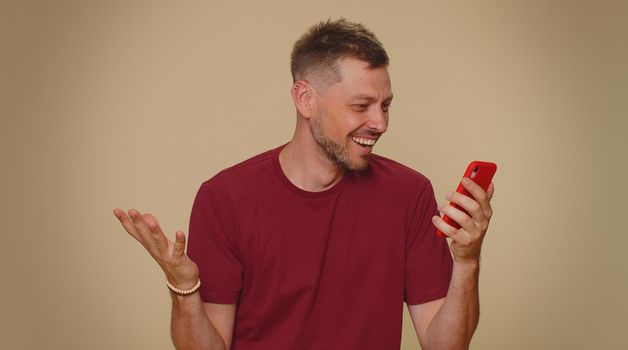 Happy excited joyful young man use smartphone typing browsing say wow yes, found out great big win news doing winner gesture. Adult stylish male guy isolated on beige studio wall background indoors