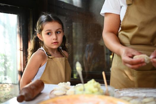 Beautiful Caucasian little girlwuth two cute ponytails, wearing a beige apron, standing near her mom in the country kitchen while cooking dumplings for the lunch. Baking concept. Homemade Vareniki