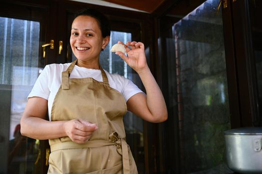 Charming happy African American woman in chef's apron, holds moldy dumplings, smiling sweetly with cheerful toothy smile, enjoys a cooking class cooking homemade Ukrainian varenyky in rustic kitchen