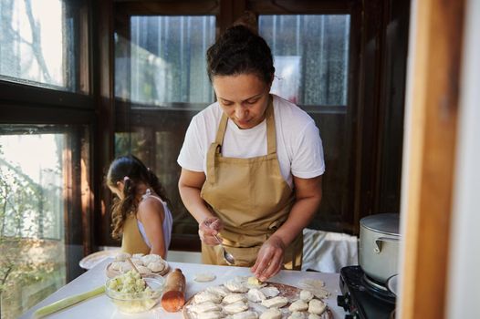 Beautiful multi-ethnic woman, a pleasant housewife in apron, puts mashed potatoes on a rolled out dough, enjoys cooking dumplings for lunch with her daughter in the country kitchen. Baking concept