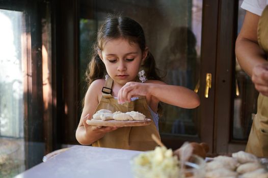 Artisanal scene of beautiful child, a little girl with two cute ponytails, wearing chef's apron, standing at kitchen table and sprinking white flour on homemade dumplings or vareniki on a wooden board