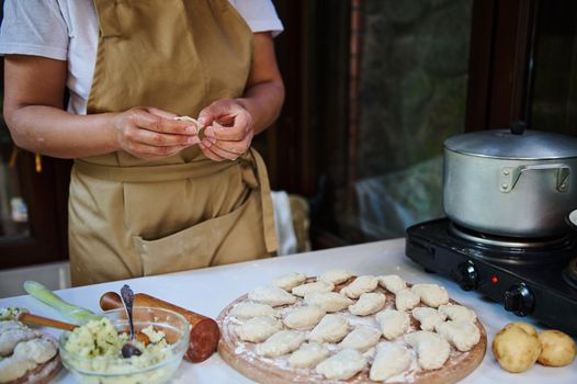 Selective focus on homemade dumplings stuffed with mashed potatoes and chopped dill on wooden board, against a housewife in beige chefs apron, cooking Ukrainian national dish for the dinner - Vareniki