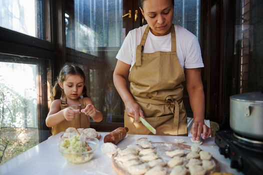 Loving mother and her adorable little daughter molding dumplings. Multi-ethnic pretty woman in a beige chef's apron, a housewife cooking Ukrainian vareniki in the rural summer kitchen. Baking concept