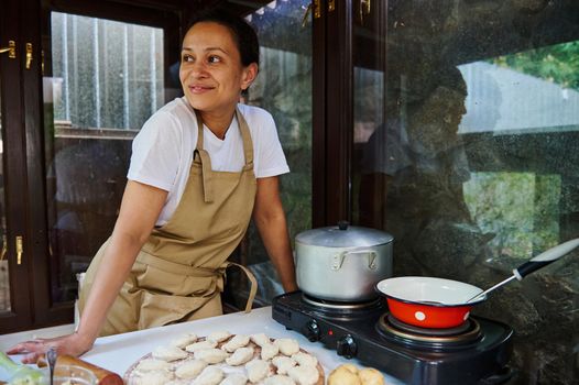 Attractive young Latin American pretty woman, pleasant housewife wearing chef's apron cooking dumplings. Ukrainian national dish homemade varenyky. Russian Plemeni. Italian Ravioli. Baking concept