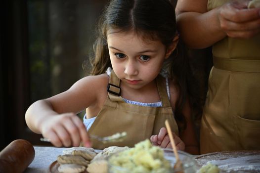 Close-up portrait of a pretty Caucasian child girl, wearing a beige chef's apron, standing by a table with dough and ingredients, helping her mother cooking Ukrainian varenyky in the country kitchen