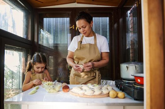 Beautiful woman and her cute little daughter kneading dough and cutting it into portions, preparing homemade dumplings, Ukrainian traditional dish- Vareniki, in the summer country kitchen