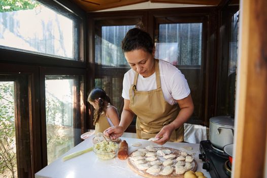 Happy multiethnic woman, young loving mother in beige chef's apron, preparing dumpling or pelmeni dough or Ukrainian varenyky, with her adorable little daughter in the country kitchen. Family pastime