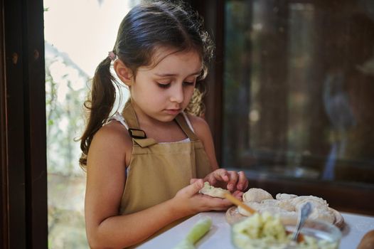 Beautiful Caucasian child, a little cook, adorable baby girl with two ponytails, in beige chef's apron stuffs mashed potatoes and molds dumplings in a country kitchen, helps her mother cooking dinner