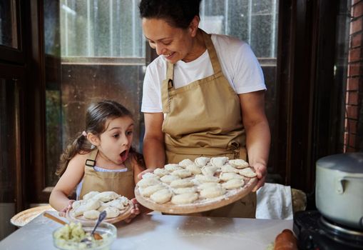 Happy multi-ethnic beautiful woman, a loving mom and daughter, both of them wearing beige chef's aprons, smiling while holding wooden boards with homemade Ukrainian vareniki sprinkled with white flour