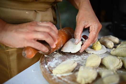 Details: Hands of a chef in beige apron, using a rolling pin, rolls out dough on a wooden board, prepares homemade dumplings or Ukrainian national dish Varenyky. Selective focus. Baking cooking class