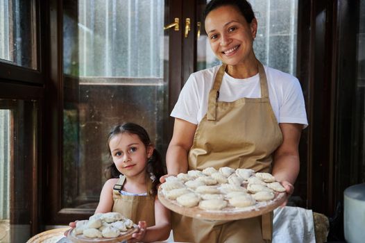 Charming multi-ethnic woman, a loving mom and daughter, both of them wearing beige chef's aprons, smiling at camera, holding wooden boards with dumplings sprinkled with white flour. Baking concept