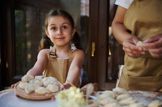 Cute child, beautiful Caucasian little girl wearing a beige chef's apron, smiling a cheerful sweet smile, holding a wooden board with homemade dumplings. Artisanal, rural scene of cooking food