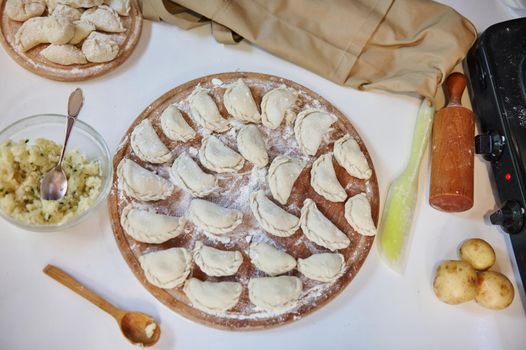 Top view. Rustic kitchen table with uncooked dumplings stuffed with mashed potatoes and sprinkled with flour on a wooden cutting board with ingredients. Traditional Ukrainian dish - Varenyky