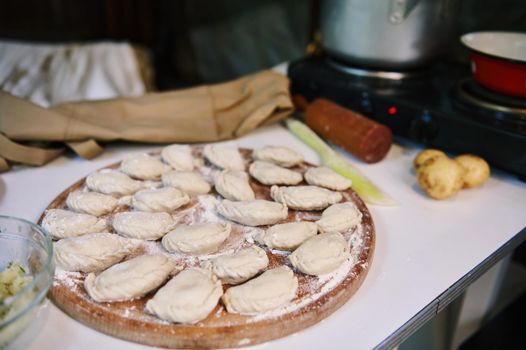 Selective focus. Close-up of homemade dumplings, Ukrainian varenyky on a wooden board, sprinkled with flour and a pot with boiling water on an electric stove in the rustic summer country kitchen