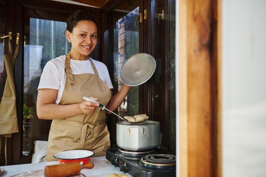 Charming multi-ethnic woman, pleasant housewife in beige chef's apron, standing by countertop, stirring homemade dumplings in the pot, preparing Ukrainian varenyky in rustic kitchen, smiling at camera