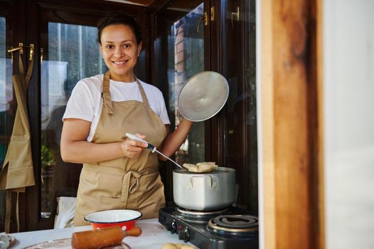 Pleasant woman, a housewife in beige chef's apron, taking out freshly brewed dumplings, vareniki, pelmeni, cute smiling at the camera. Homemade traditional Ukrainian or Russian culinary and traditions