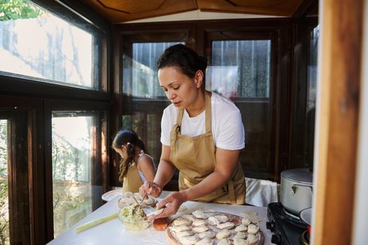 Pretty woman putting mashed potatoes on a rolled out dough and enjoying cooking dumplings with her cute daughter. Pretty little girl learns making vareniki, using rolling pin rolls out the dough