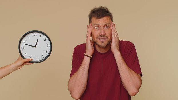 Confused young man in red t-shirt with anxiety checking time on clock, running late to work, being in delay, deadline. Adult stylish male guy looking at hour, minutes, worrying to be punctual, indoors